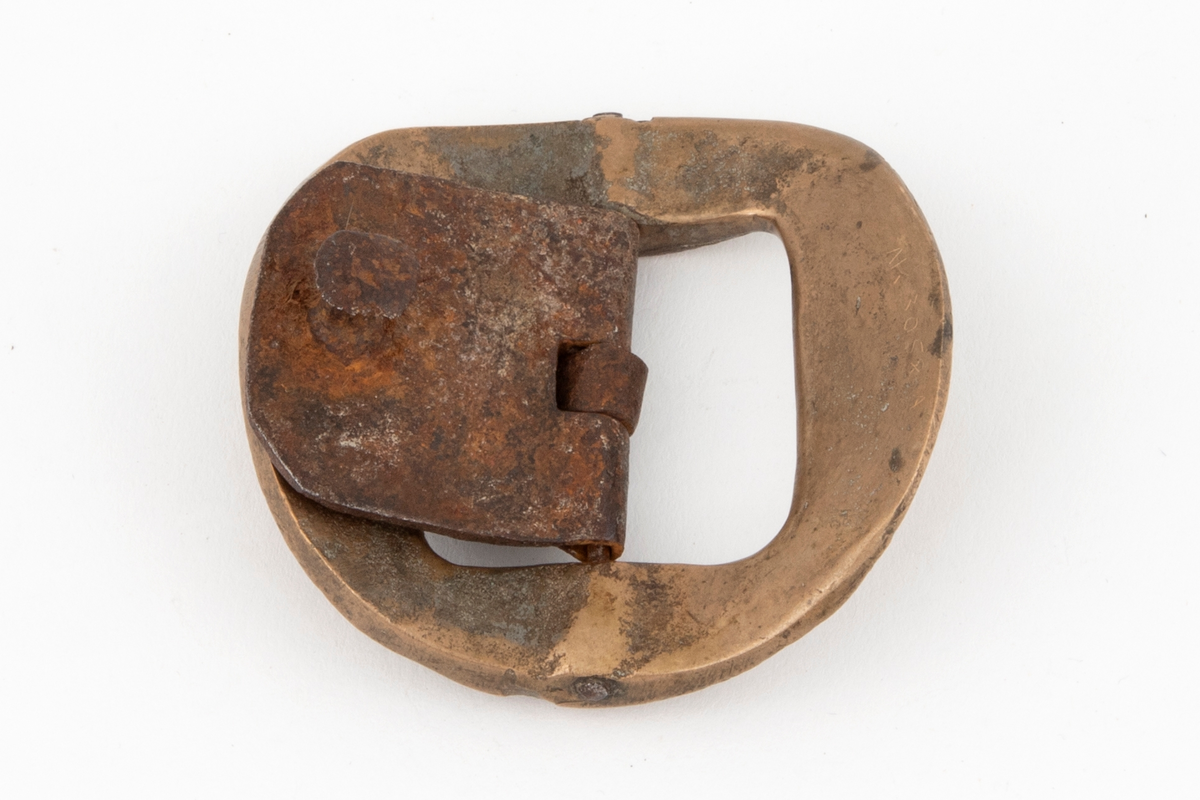 Skospenner av bronse støpt i krummet, nesten rund form med indre firkantet åpning. På to av rammesidene siselert rosett og volutter. De to andre sidene har båndlignende ornament. Stolpe med torn og feste i form av tunge med knapp i jern.