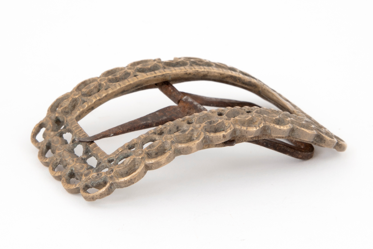 Skospenner av bronse støpt i krummet, rektangulær form med avrundede hjørner. Indre rektangulær åpning med skarpe hjørner. Rammen er gjennombrutt med dobbelt rad motstilte buer. Mellom buene bord med rombiske og runde groper. Stolpe med torn og feste med liten fast torn i jern.