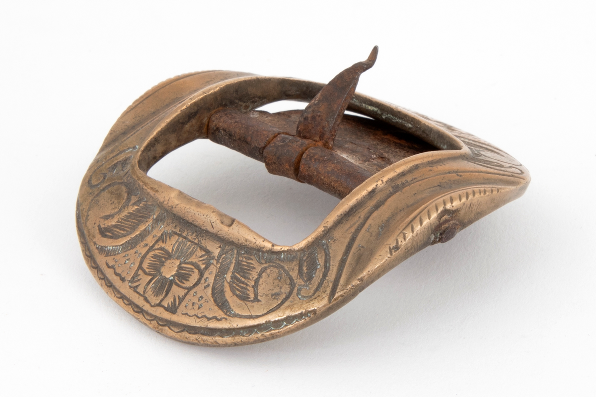 Skospenner av bronse støpt i krummet, nesten rund form med indre firkantet åpning. På to av rammesidene siselert rosett og volutter. De to andre sidene har båndlignende ornament. Stolpe med torn og feste i form av tunge med knapp i jern.