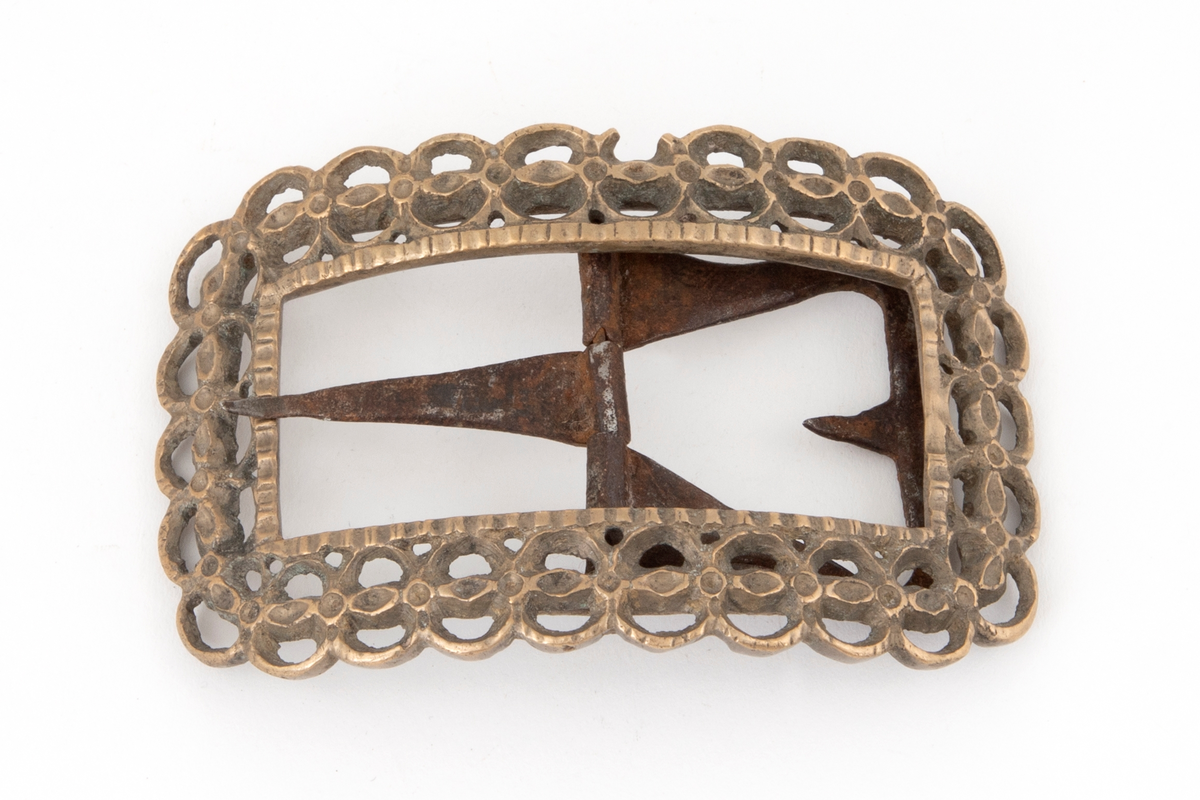 Skospenner av bronse støpt i krummet, rektangulær form med avrundede hjørner. Indre rektangulær åpning med skarpe hjørner. Rammen er gjennombrutt med dobbelt rad motstilte buer. Mellom buene bord med rombiske og runde groper. Stolpe med torn og feste med liten fast torn i jern.