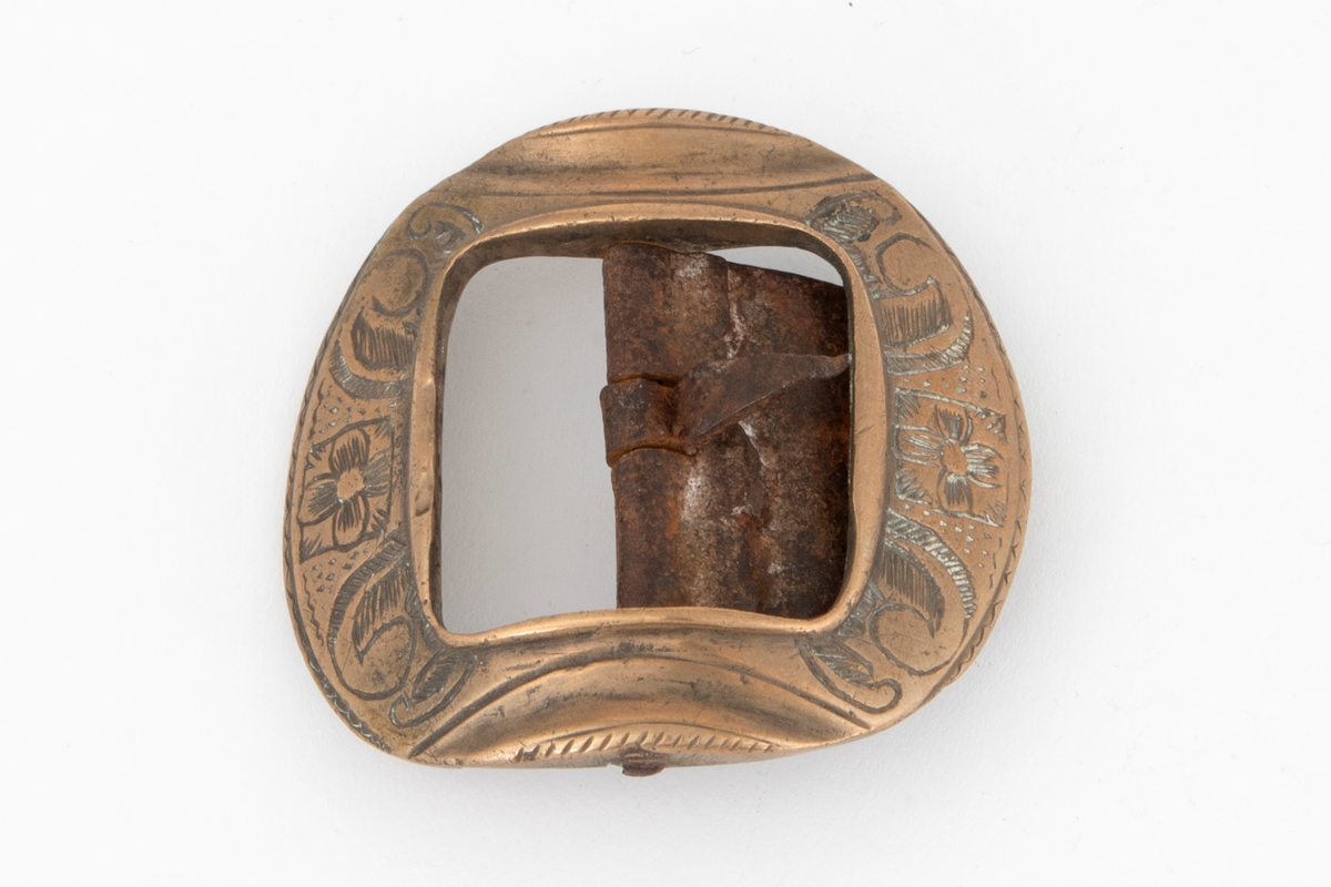 Skospenner av bronse støpt i krummet, nesten rund form med indre firkantet åpning. På to av rammesidene siselert rosett og volutter. De to andre sidene har båndlignende ornament. Stolpe med torn og feste i form av tunge med knapp i jern.