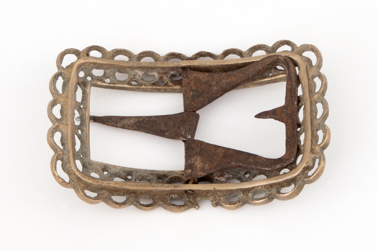 Skospenner av bronse støpt i krummet, rektangulær form med avrundede hjørner. Indre rektangulær åpning med skarpe hjørner. Rammen er gjennombrutt med dobbelt rad motstilte buer. Mellom buene bord med rombiske og runde groper. Stolpe med torn og feste med liten fast torn i jern.