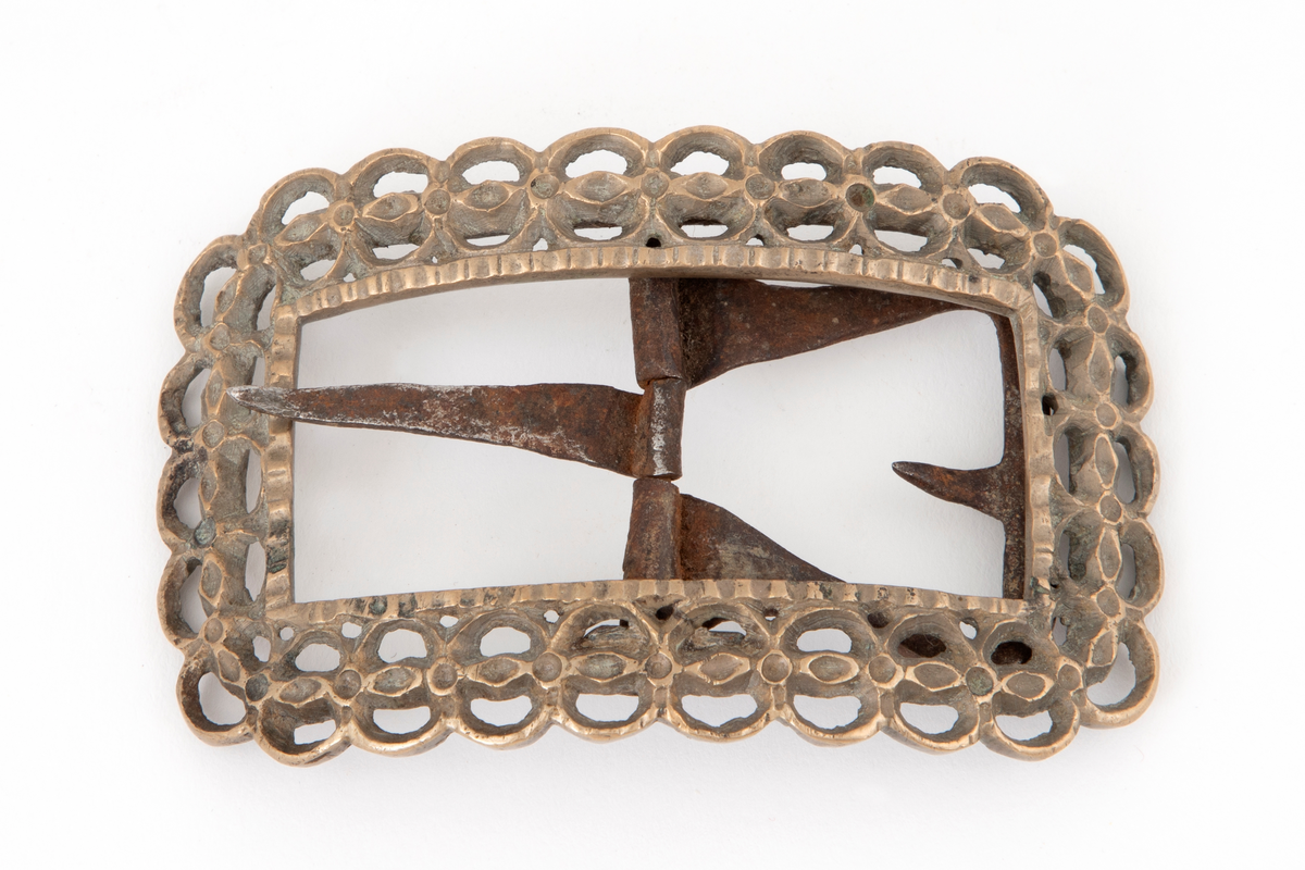 Skospenner av bronse støpt i krummet, rektangulær form med avrundede hjørner. Indre rektangulær åpning med skarpe hjørner. Rammen er gjennombrutt med dobbelt rad motstilte buer. Mellom buene bord med rombiske og runde groper. Stolpe med torn og feste med liten fast torn i jern.