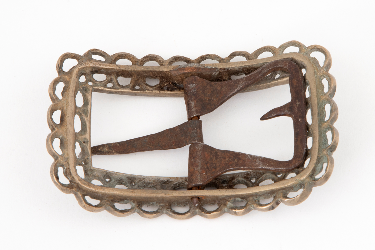 Skospenner av bronse støpt i krummet, rektangulær form med avrundede hjørner. Indre rektangulær åpning med skarpe hjørner. Rammen er gjennombrutt med dobbelt rad motstilte buer. Mellom buene bord med rombiske og runde groper. Stolpe med torn og feste med liten fast torn i jern.