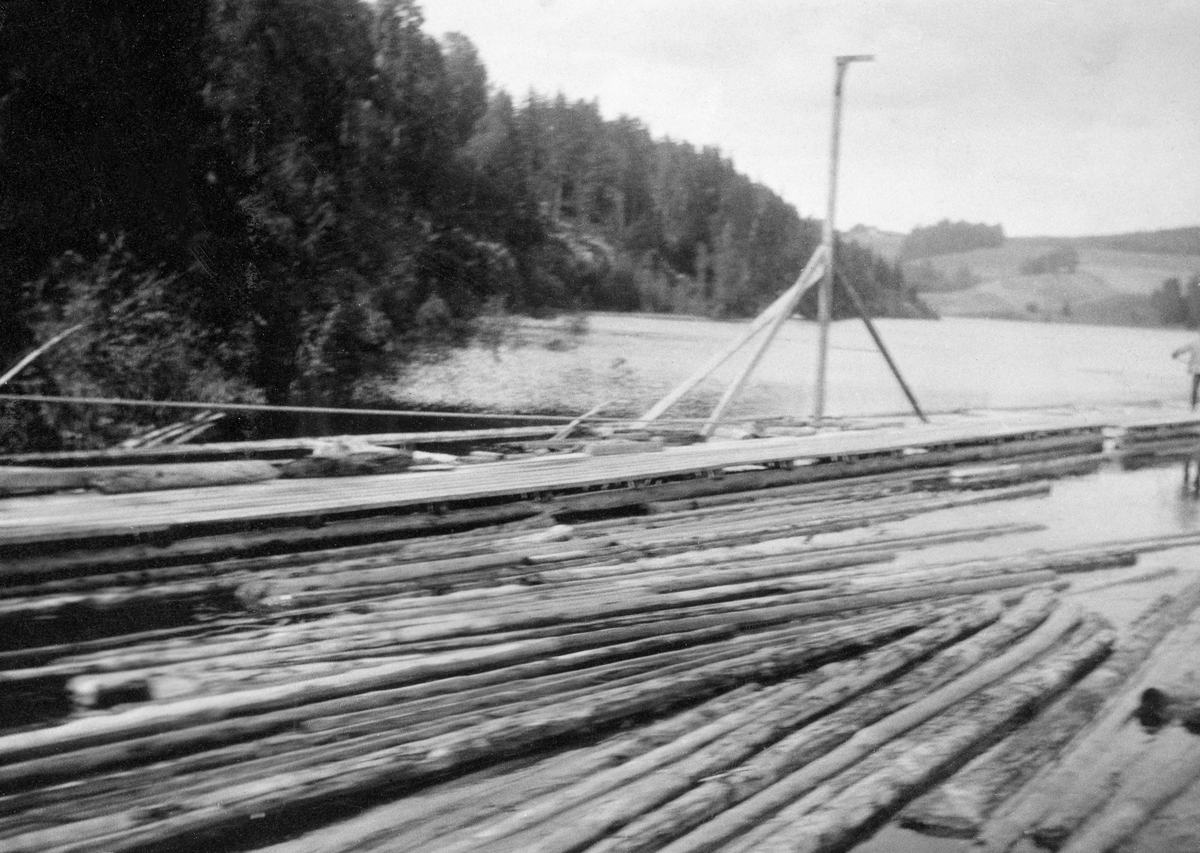 Fra den indre delen av «påstikkingslensa» ved Solbergfoss kraftstasjon i Askim (nedre Glomma).  Fotografiet er tatt fra en av flåtegangene (lensearmene) mot den andre, antakelig i den indre sonen mot inntaket til tømmerrenna, der lensa var smal.  På vannflata i forgrunnen ligger det en del barket fløtingstømmer med lengderetning som er noenlunde parallell med strømretningen i renna stokkene snart skulle over i.  På motsatt side av ser vi en lensearm som er sammenkjedet av flytende pongtonger med plankegolv og med tømmer i understellet.  I bakkant av flåtegangene er det montert lyktestolper med elektriske lamper.  Stolpene holdes i vertikal posisjon av tre skråstivere (side- og bakovervendte).  Den elektriske belysningen var viktig for at fløterne skulle kunne arbeide lange dager med påstikning her, også utover høsten. Her i den nedre delen av vassdraget kunne dette arbeidet i prinsippet fortsette fram til om lag 1. november, og da begynte dagslysperiodene å bli korte.  I bakgrunnen ser vi (litt uskarpt) hvordan kraftverksdammens strender møter det forholdsvis bratte Holmsberget, hvor det da fotografiet ble tatt vokste litt glissen skog.