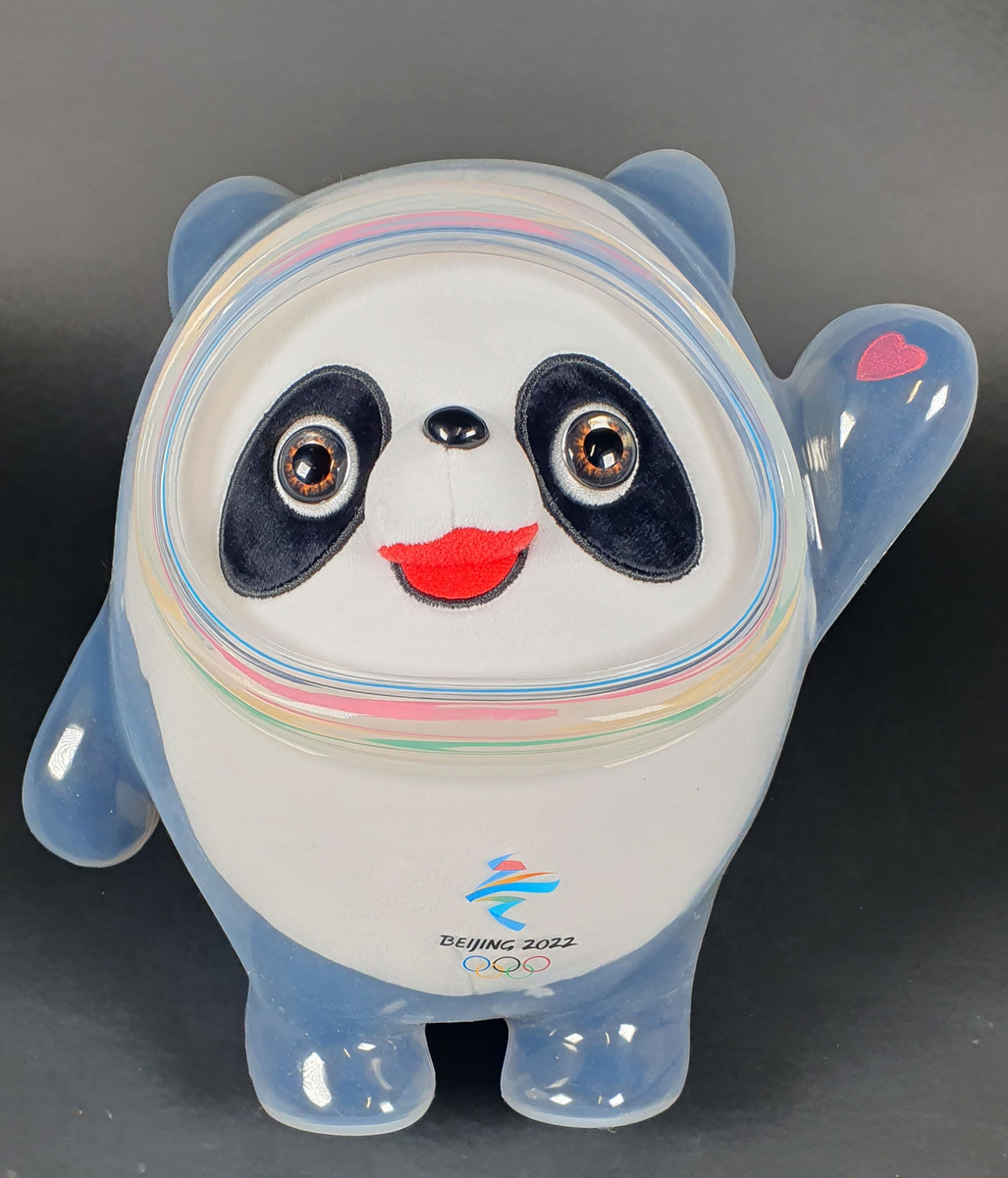 Hvit og svart panda-bamse, med knapper til øyne og et rødt hjerte på venstre hånd. Kledd i en transparent plastikkdrakt. Maskot fra vinter-OL i Beijing 2022, med emblem midt foran.