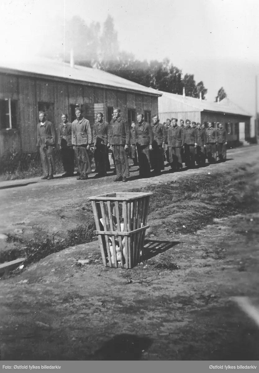 AT-leir  (arbeidstjeneste) 2. verdenskrig ca.1943. Leiren lå der Varteig skole ligger i dag. 
Gruppe har tatt oppstilling til morgenrevelje. Brakker i bakgrunnen.