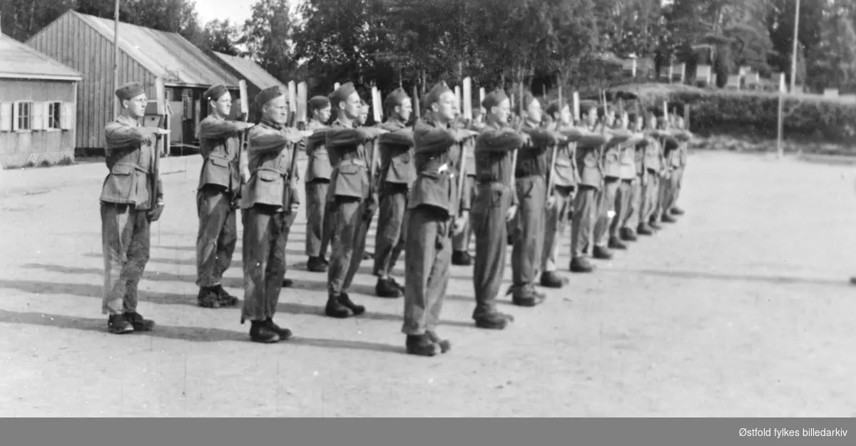 AT-leir  (arbeidstjeneste) 2. verdenskrig ca.1943. Leiren lå der Varteig skole ligger i dag. 
Gruppe har tatt oppstilling klar til eksersis med spade på skuldra. Brakker i bakgrunnen.