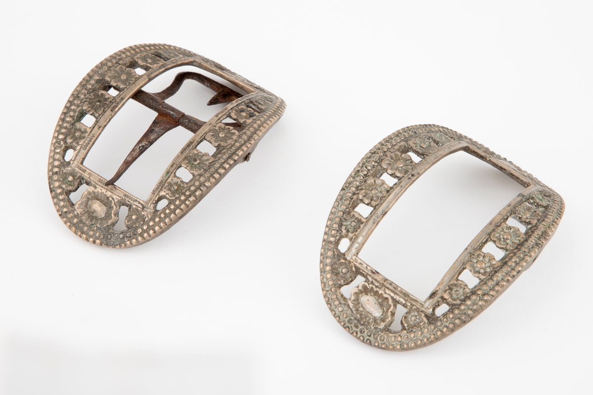 Skospenner støpt i krummet, oval form. Indre rektangulær åpning med glatt ramme. Mellom denne og den ytterste raden som har to perlerader, er et gjennombrutt parti med en rekke rosetter i forskjellige størrelser. Stolpe med torn og feste med liten fast torn i jern på den ene spennen. Den andre mangler stolpe og feste.