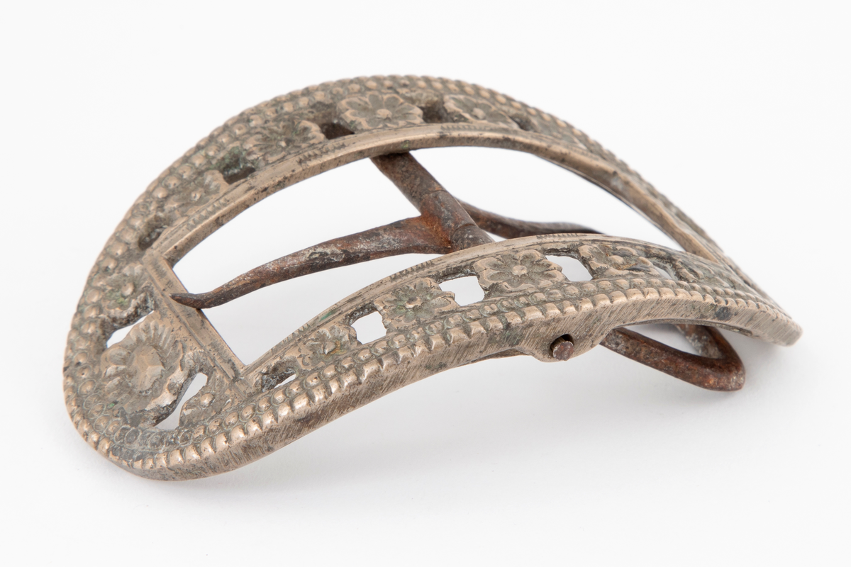 Skospenner støpt i krummet, oval form. Indre rektangulær åpning med glatt ramme. Mellom denne og den ytterste raden som har to perlerader, er et gjennombrutt parti med en rekke rosetter i forskjellige størrelser. Stolpe med torn og feste med liten fast torn i jern på den ene spennen. Den andre mangler stolpe og feste.