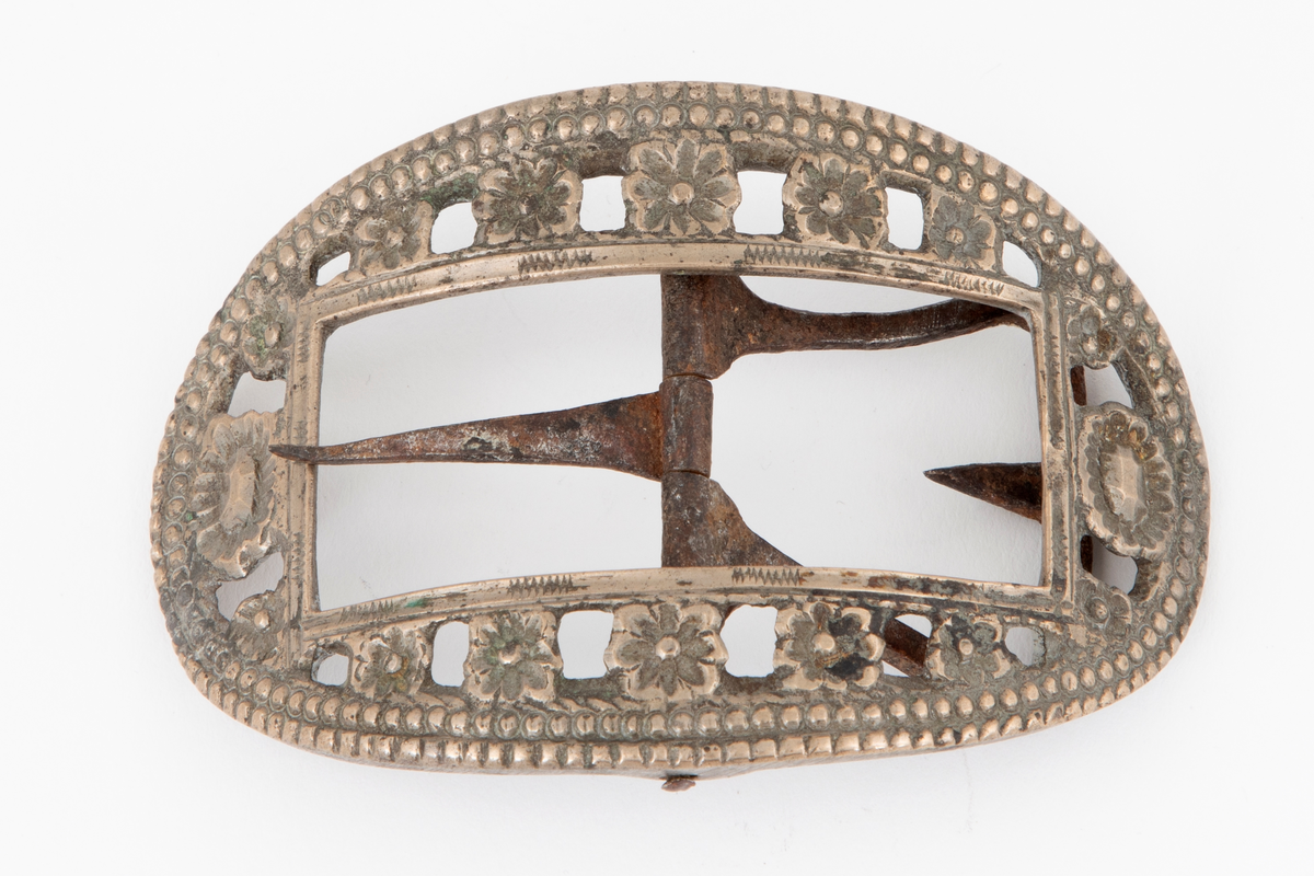 Skospenner støpt i krummet, oval form. Indre rektangulær åpning med glatt ramme. Mellom denne og den ytterste raden som har to perlerader, er et gjennombrutt parti med en rekke rosetter i forskjellige størrelser. Stolpe med torn og feste med liten fast torn i jern på den ene spennen. Den andre mangler stolpe og feste.