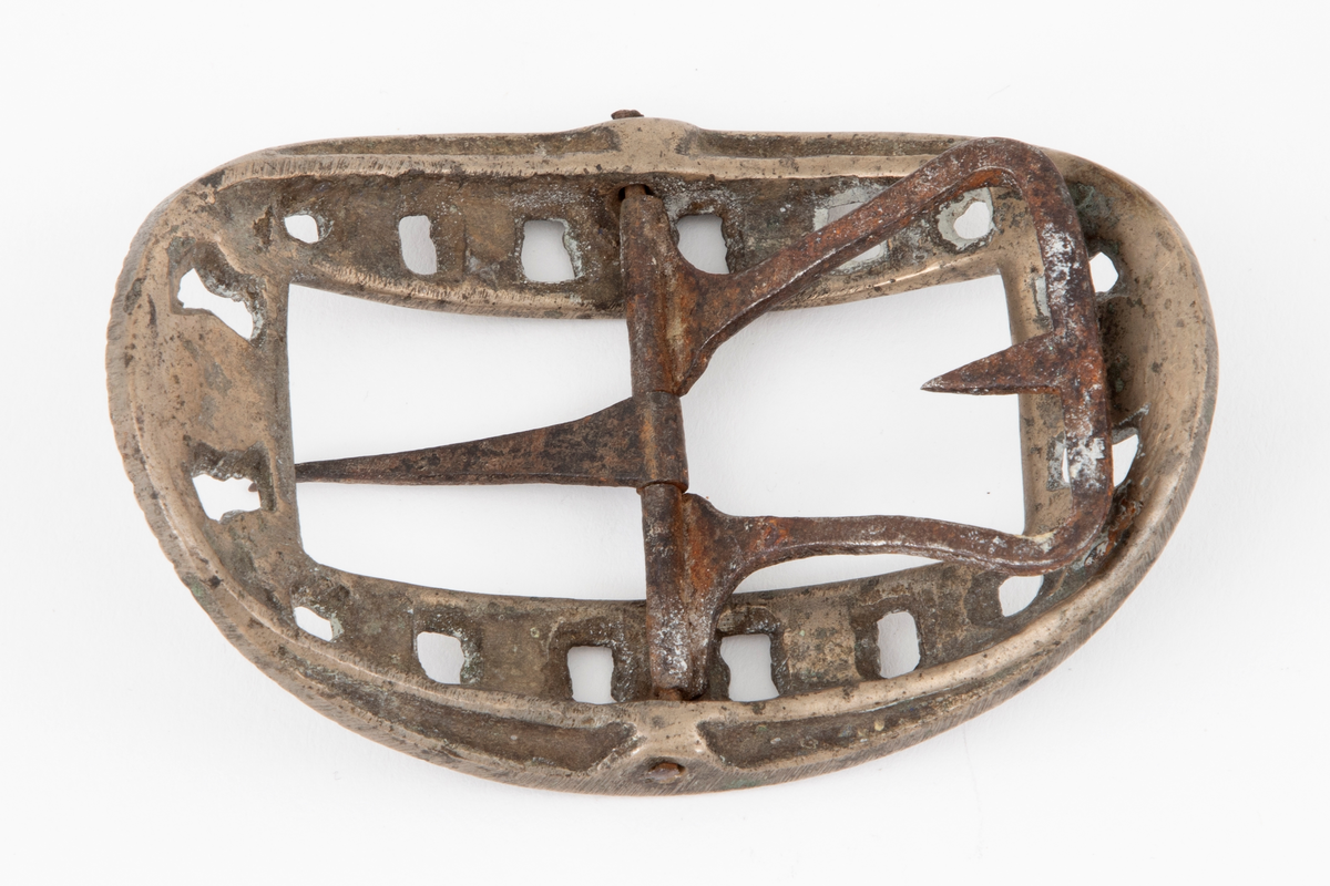 Skospenner støpt i krummet, oval form. Indre rektangulær åpning med glatt ramme. Mellom denne og den ytterste raden som har to perlerader, er et gjennombrutt parti med en rekke rosetter i forskjellige størrelser. Stolpe med torn og feste med liten fast torn i jern på den ene spennen. Den andre mangler stolpe og feste.