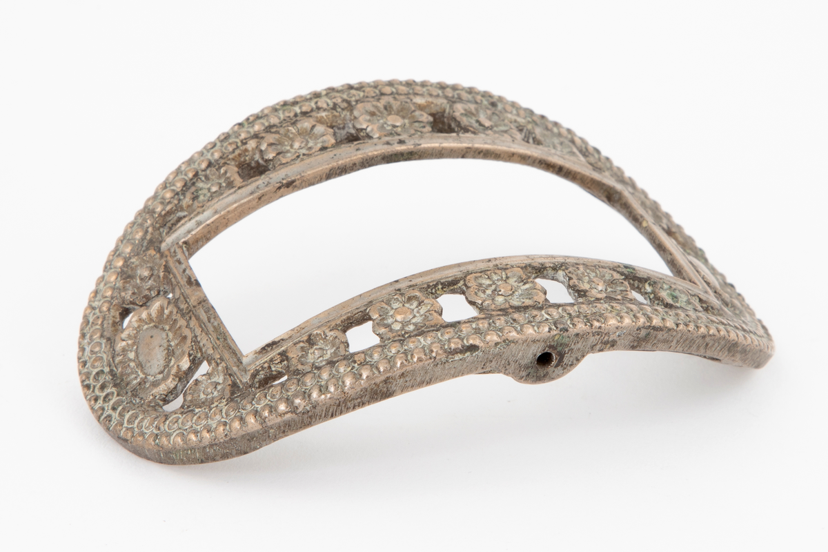 Skospenner støpt i krummet, oval form. Indre rektangulær åpning med glatt ramme. Mellom denne og den ytterste raden som har to perlerader, er et gjennombrutt parti med en rekke rosetter i forskjellige størrelser. Stolpe med torn og feste med liten fast torn i jern på den ene spennen. Den andre mangler stolpe og feste.