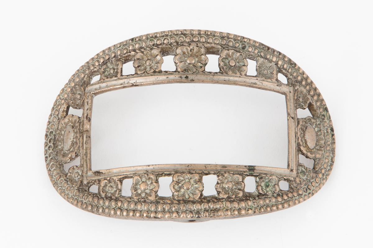 Skospenner støpt i krummet, oval form. Indre rektangulær åpning med glatt ramme. Mellom denne og den ytterste raden som har to perlerader, er et gjennombrutt parti med en rekke rosetter i forskjellige størrelser. Stolpe med torn og feste med liten fast torn i jern på den ene spennen. Den andre mangler stolpe og feste.