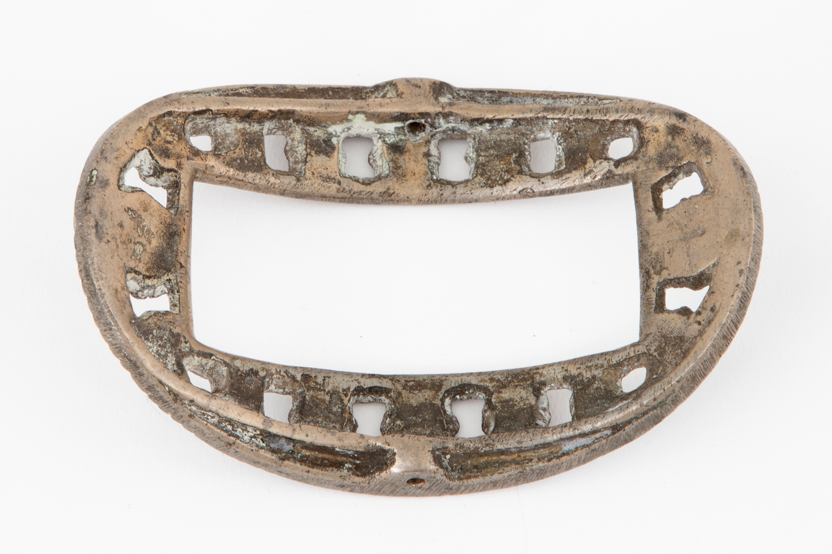 Skospenner støpt i krummet, oval form. Indre rektangulær åpning med glatt ramme. Mellom denne og den ytterste raden som har to perlerader, er et gjennombrutt parti med en rekke rosetter i forskjellige størrelser. Stolpe med torn og feste med liten fast torn i jern på den ene spennen. Den andre mangler stolpe og feste.