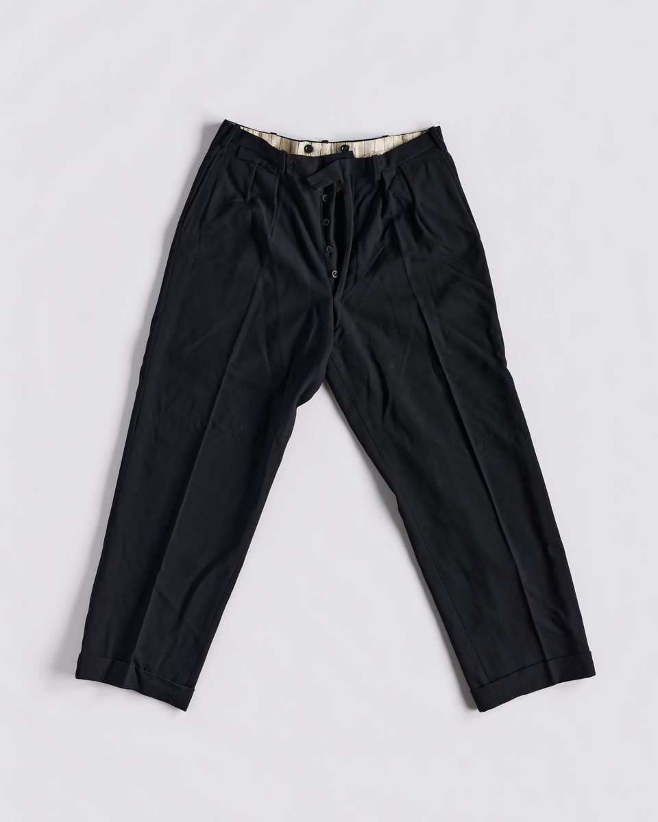 Dressbukse i bomull. Gylfen lukkes med 6 knapper og en hekter. To baklommer og to stikklommer foran. Ved linningen er det en liten lomme med klaff. Smygstoler i linningen til belte. Knapper på innsiden av linningen til bukseseler. Brett nederst på buksebeina.