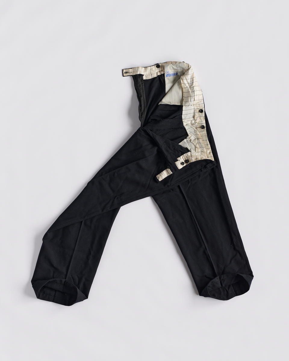 Dressbukse i bomull. Gylfen lukkes med 6 knapper og en hekter. To baklommer og to stikklommer foran. Ved linningen er det en liten lomme med klaff. Smygstoler i linningen til belte. Knapper på innsiden av linningen til bukseseler. Brett nederst på buksebeina.