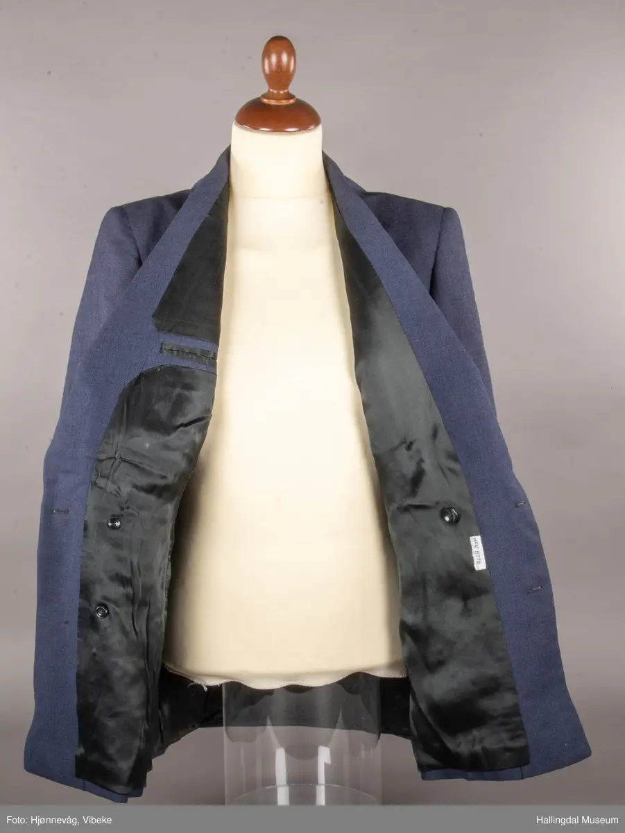 Blågrå dressjakke med slag og dobbel rad med knapper, 3 knapper på hver side. En brystlomme på venstre side, 2 jakkelommer uten klaff. 3 knapper framme på hvert erme. Foret med svart tøy av kunstsilke. En liten innerlomme. Jakka har antagelig tilhørt skredder Odd Navrud.