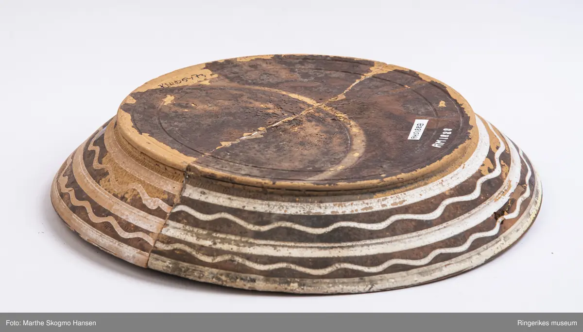 Brunglassert keramikkfat med hvit dekor i form av to rette striper og to bølgete striper på utsiden. Linje med prikker og bladornament på innsiden. Fatet er sprukket i to deler, men limt sammen igjen