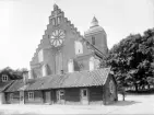 Den pampiga östfasaden döljer från fotografens perspektiv närmast den i övrigt storslagna Vårfrukyrkan i Skänninge. Kyrkan uppfördes efter tyskt mönster och invigdes i början av 1300-talet, helgad åt jungfru Maria som givit kyrkan sitt namn. För det låga trähuset i förgrunden kan inga tillfredsställande svar ges inom ramen för uppgiften. På gaveln annonseras 