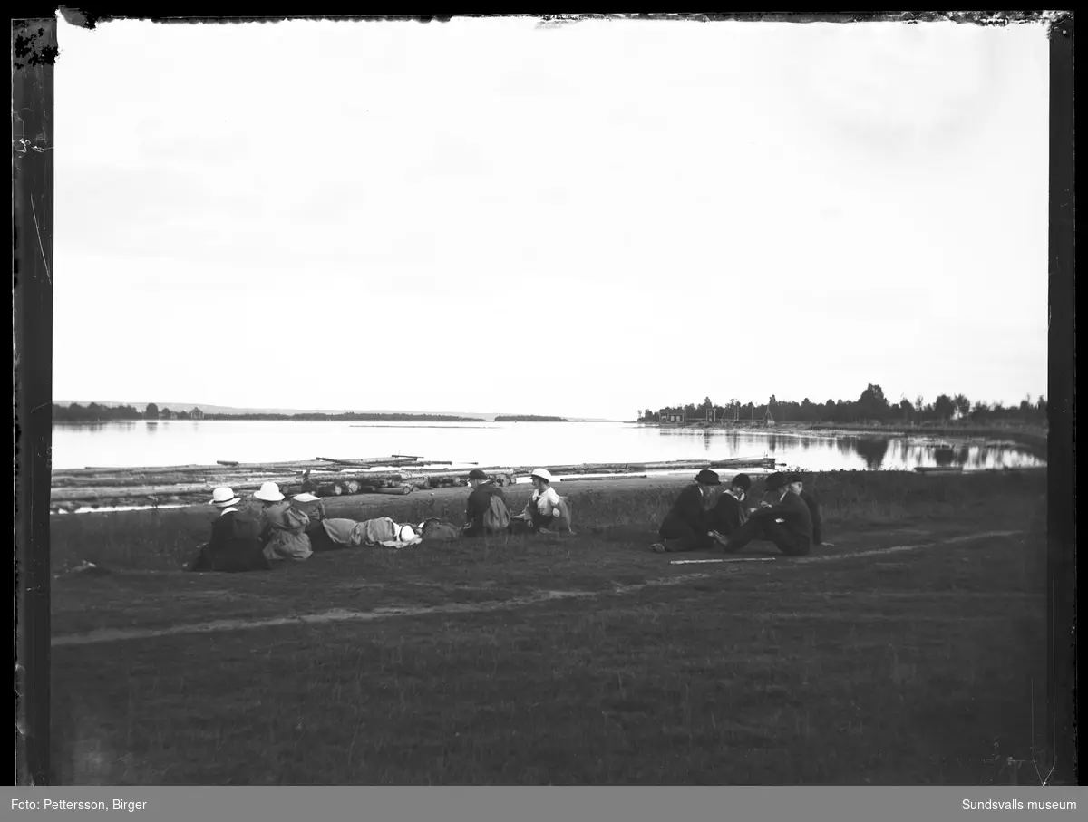 Yngre män och kvinnor sitter i gräset vid strandkanten i skärgårdsmiljö.