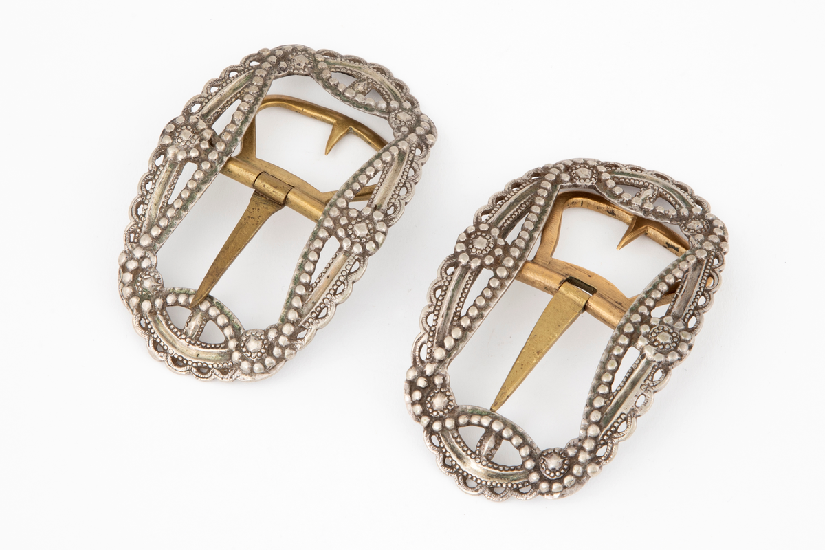 Skospenner støpt i krummet, oval form. Siselert og med gjennombrutt dekor i relieff med rosetter, festons, perlerad og ytterst gjennombrutt tunget rand. Stolpe med torn og feste med liten torn i messing.