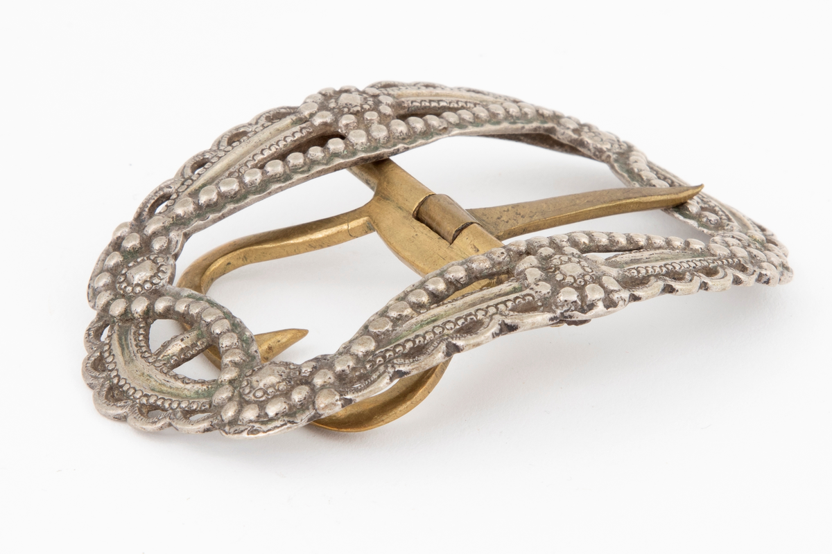 Skospenner støpt i krummet, oval form. Siselert og med gjennombrutt dekor i relieff med rosetter, festons, perlerad og ytterst gjennombrutt tunget rand. Stolpe med torn og feste med liten torn i messing.