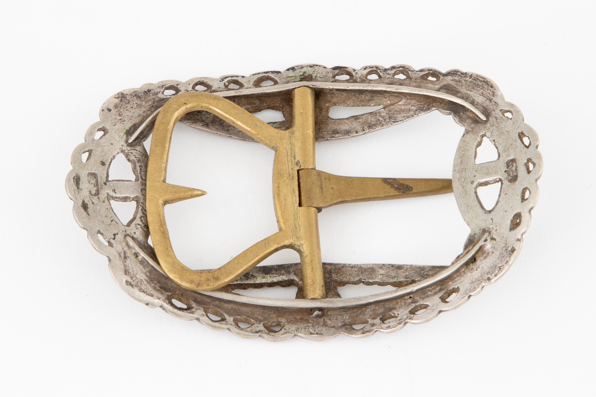 Skospenner støpt i krummet, oval form. Siselert og med gjennombrutt dekor i relieff med rosetter, festons, perlerad og ytterst gjennombrutt tunget rand. Stolpe med torn og feste med liten torn i messing.