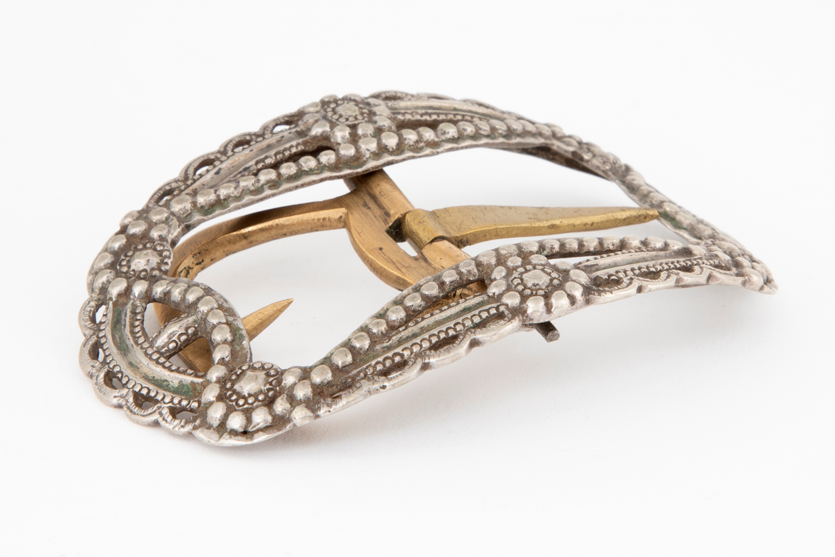 Skospenner støpt i krummet, oval form. Siselert og med gjennombrutt dekor i relieff med rosetter, festons, perlerad og ytterst gjennombrutt tunget rand. Stolpe med torn og feste med liten torn i messing.