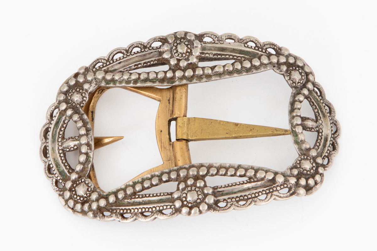 Skospenner støpt i krummet, oval form. Siselert og med gjennombrutt dekor i relieff med rosetter, festons, perlerad og ytterst gjennombrutt tunget rand. Stolpe med torn og feste med liten torn i messing.