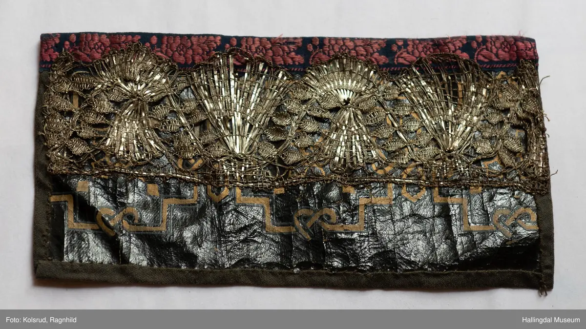 Voksduk med mønster i gull og grått. Påsatt bred sølvknipling. Øverst blomsterete bånd. Foret med sort tøy. Tråklesting i sort og hvitt.