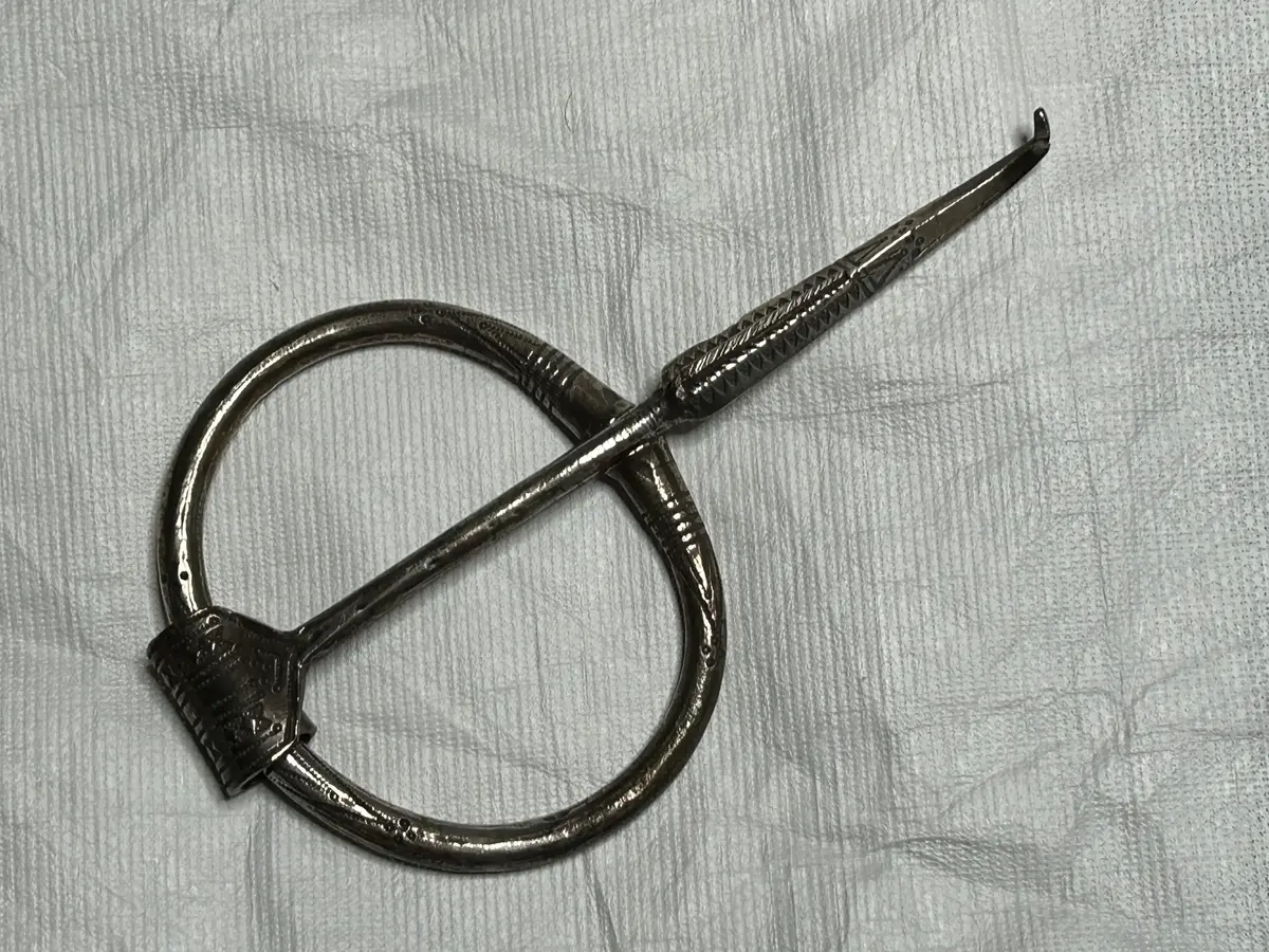 AV SILVER; RINGEN OVAL MED RUND GENOMSKÄRNING, VIDGANDE SIG MOT ÄNDARNA, DEKORERAD MED INPUNSADE PUNKTER, BILDANDE DUBBLA, SPETSIGT TRIANGULÄRA MÖNSTER MED TRIANGLARNAS BASER MOT HALVA RINGEN OMSLUTANDE PARALLELLA CIRKLAR. NÅLEN UPPTILL NÄRMAST RUND, NEDTILL ROMBISK, AVSMALNANDE MOT, (DEN NUMERA BÖJDA) SPETSEN, DEN KRING RINGEN RULLADE ÄNDEN BRED, PLATT. DEKORERAD PÅ ÖVERSIDAN MED INPUNSADE ROMBER OCH PUNKTER, BILDANDE STRECK OCH TRIANGLAR. NÅLENS L. 15,5, RINGENS DIAM. 8,8 - 7,7 CM. FRÅN KARLS 1'10 I TINGSTÄDE. VER. 566/66.