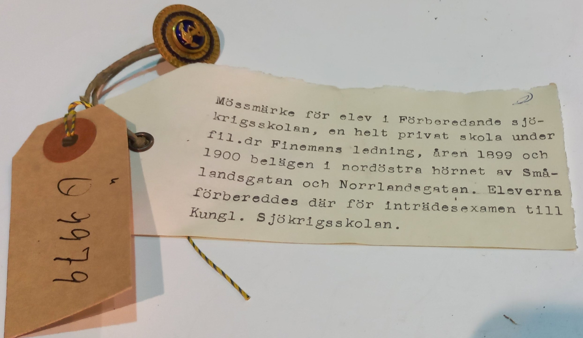 Mössmärke, runt, förgylld kokard försett med ett runtom gående blått band. Å kokarden är fastsatt en välvd knapp av blå emalj i vars mitt ett ankare är fastsatt. Användes som mössmärke för elever i "Förberedande sjökrigsskolan" - en helt privat skola 1891-1904, under fil dr Finemans ledning, åren 1894-95 och 1896-1901. Skolan var belägen i NÖ hörnet av Smålandsgatan och Norrlandsgatan. Eleverna förbereddes där för inträdesexamen till Kungl Sjökrigsskolan..