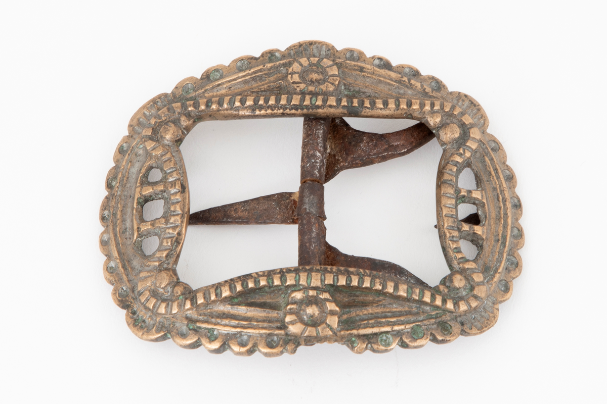 Skospenner støpt i krummet, oval form med innvendig firkantet åpning med buede sider (festons). Delvis gjennombrutt og dekor i relieff, to rosetter og festons med rifler. Ytre rand tunget. Stolpe med torn og feste med liten fast torn av jern.