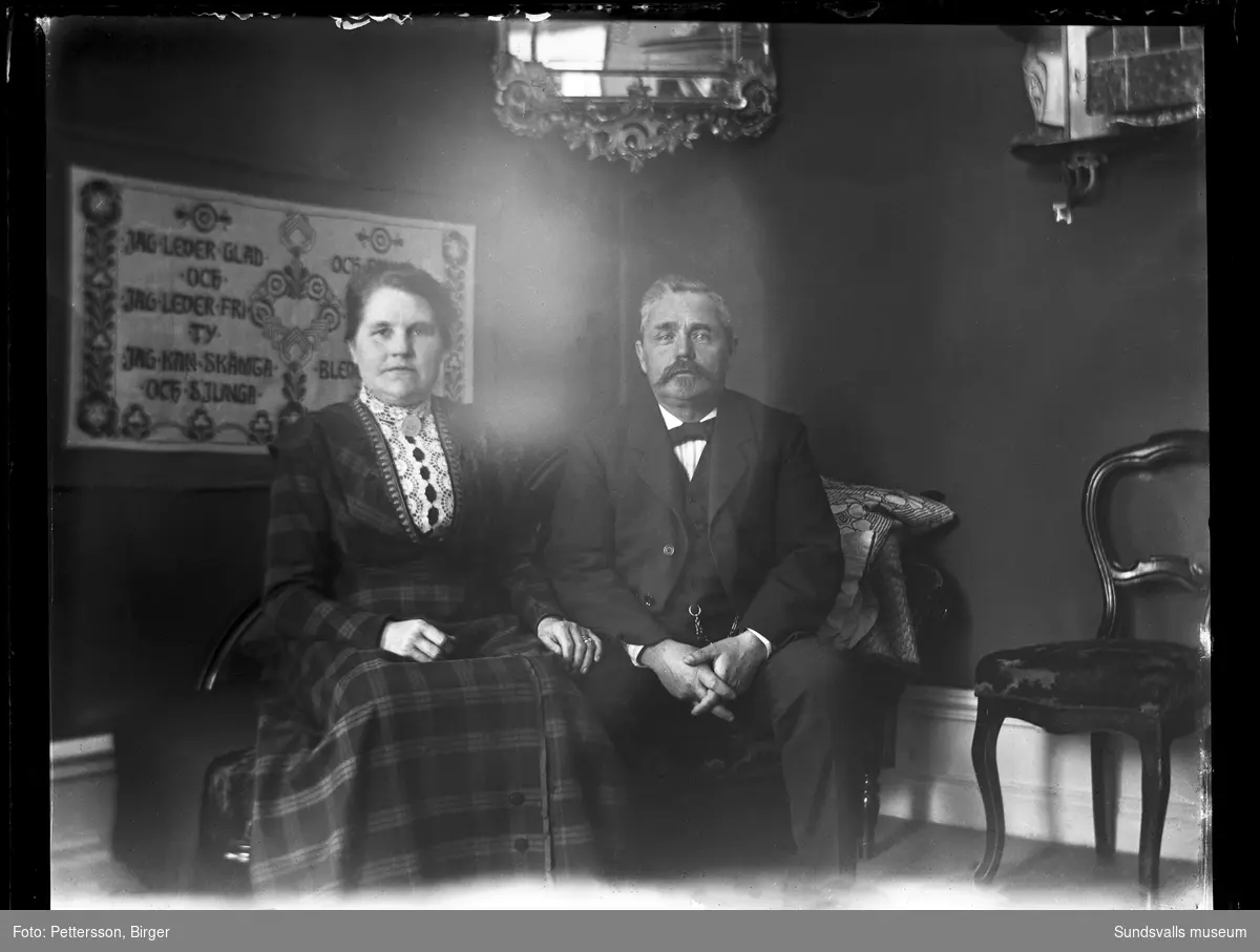 Porträttbild av ett par som sitter i en kanapé. På väggen bakom hänger en bonad med broderade visdomsord. Kvinnan är med på en bild som togs vid samma tillfälle och har nummer SuM-foto035271.