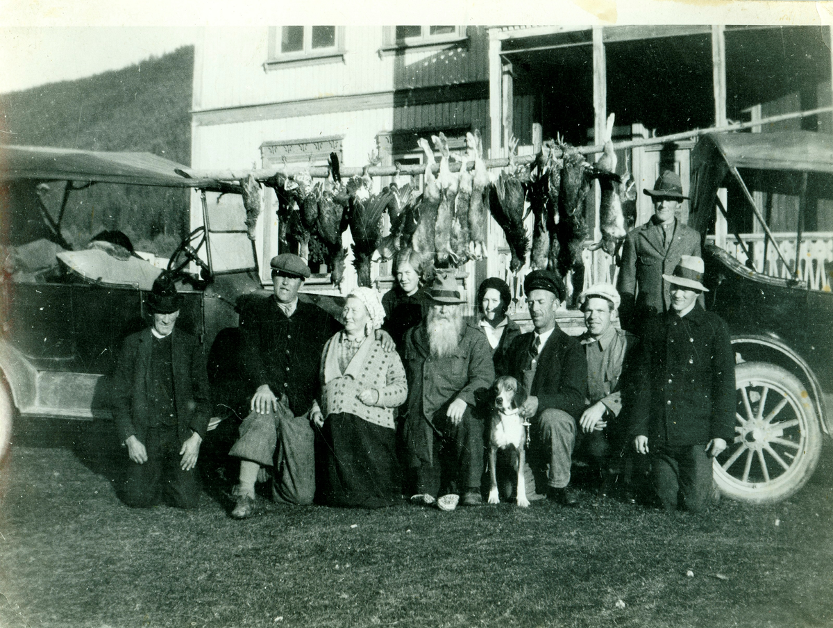 Gruppe framfor bilar og hus. Bak heng det jaktutbytte med harar og tiur. 
Frå v. Torstein Finnesgarden Gutigard, Olvar Sand, Ingebjør Bjella Gutigard, Laura Gutigard, Rosendal, ukjent,  Edvard Bø, Kristian Langsø og Olav Gutigard. Bak står Tolleiv Gutigård. Den bakerste bilen er ein Ford T 1923-25.