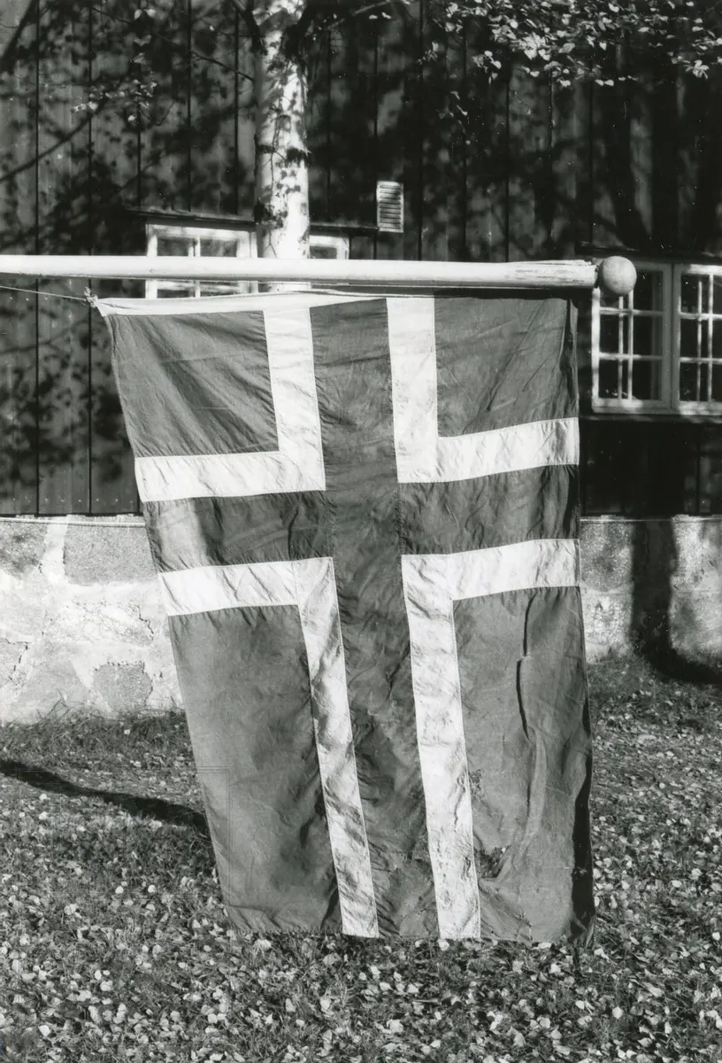 Norsk flagg. Mange hull, laget av kuer da båten lå ved fjorden. Kulen og festet på stangen er ny. Snøret er også nytt. Snøret er vanlig snor. Stangen er malt hvit.