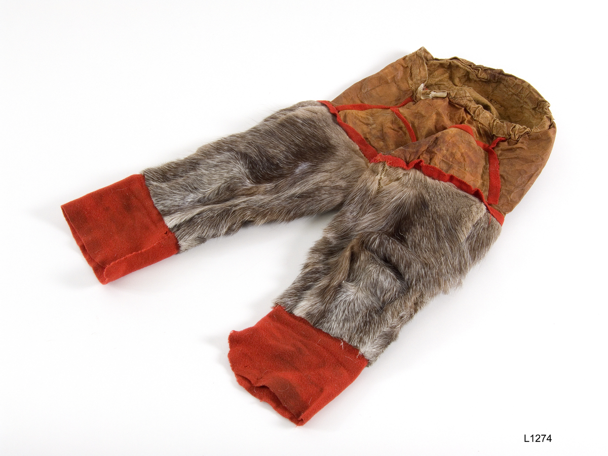 Guttebukse. Lengde fra koftelinning til nedre kant 44 cm . Grått reinskinn med bred, rød filt kant nederst, selve broka av semsket skinn. Sømmene er pyntet med røde filtkanter. (Tilh. 1273) Forarbeidet av Marit Sara Eira . Innkjøpt hos henne av Vorren våren 1964 . Kautokeino, Finnmark. Reindriftssamer.