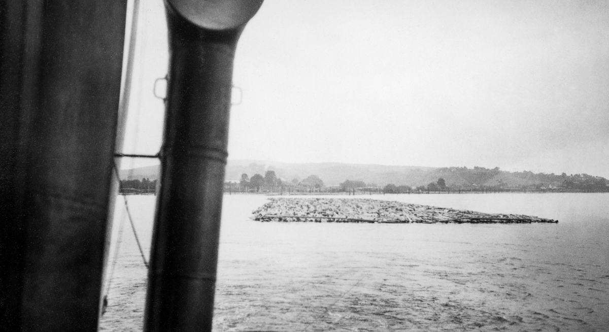 Sleping av tømmerbom, antakelig på innsjøen Øyeren i Akershus i 1925.  Fotografiet er tatt over akterdekket på en av Christiania Tømmerdirektions slepebåter.  Vi ser (til venstre i bildeflata) en skorsteinsliknende blikklur og vi ser litt av relingen bakerst i båten med slepetrossa mot tømmerbommen, som ligger et godt stykke bak båten.  Helt i bakgrunnen skimter vi et lenseanlegg inne ved land. 