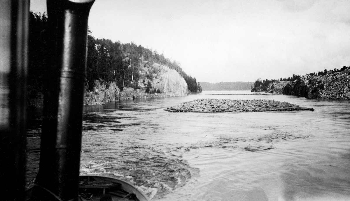 Sleping av tømmerbom ved Mørkfoss, ved innløpet fra innsjøen Øyeren til den nedre delen av Glommavassdraget.  Fotografiet er tatt over akterenden på en av Christiania Tømmerdirektions slepebåter mot det bakenforhengende tømmerslepet. Til venstre i bildet ser vi en slags skorsteinsliknende stållur som kommer opp fra dekket. Bakenfor får vi et glimt av relingen akterut i båten, med trossa slepet var forankret i. Like bak båten var vannet noe opprørt på grunn av propellaktiviteten. Slepet, som hadde en tilnærmet rektangulær form, gikk noen titalls meter bak båten, mellom forholdsvis steile, bergrike elvebredder, men med god klaring på begge sider.