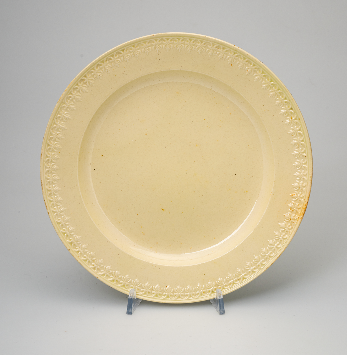 En flat tallerken av glasert steintøy (creamware). Den er hvitgul på farge (ensfarget). Flaten på midten er 16,5cm i diameter. Langs kanten er det tungekant. Innenfor tungekanten er det støpt dekor i form av en hjertebord med stiliserte blomster/bladverk. På undersiden er det dypstempel i form av fire små ruter/prikker i klase.