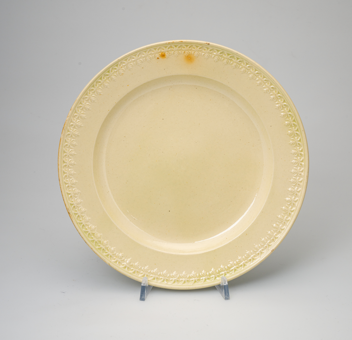 En flat tallerken av glasert steintøy (creamware). Den er hvitgul på farge (ensfarget). Flaten på midten er 16,5cm i diameter. Langs kanten er det tungekant. Innenfor tungekanten er det støpt dekor i form av en hjertebord med stiliserte blomster/bladverk. På undersiden er det dypstempel i form av fire små ruter/prikker i klase.
