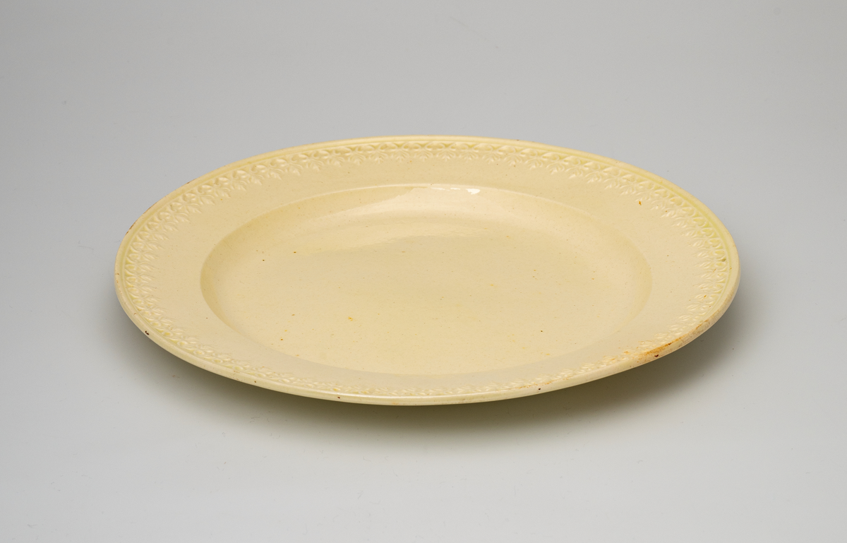 En flat tallerken av glasert steintøy (creamware). Den er hvitgul på farge (ensfarget). Flaten på midten er 16,5cm i diameter. Langs kanten er det tungekant. Innenfor tungekanten er det støpt dekor i form av en hjertebord med stiliserte blomster/bladverk. På undersiden er det dypstempel i form av fire små ruter/prikker i klase.