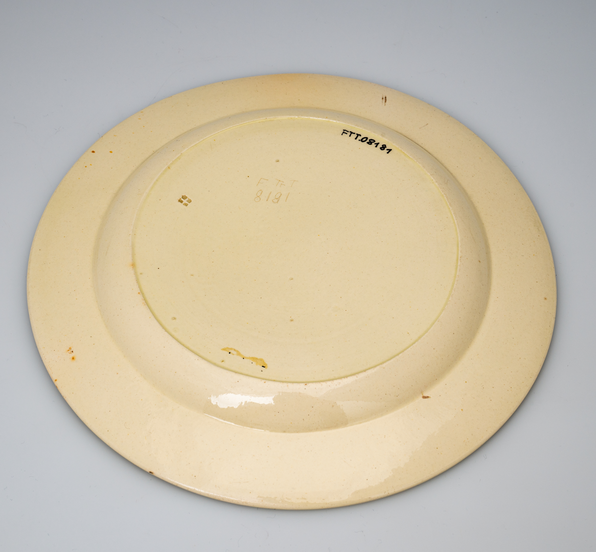 En flat tallerken av glasert steintøy (creamware). Den er hvitgul på farge (ensfarget). Flaten på midten er 16,5cm i diameter. Langs kanten er det tungekant. Innenfor tungekanten er det støpt dekor i form av en hjertebord med stiliserte blomster/bladverk. På undersiden er det dypstempel i form av fire små ruter/prikker i klase.