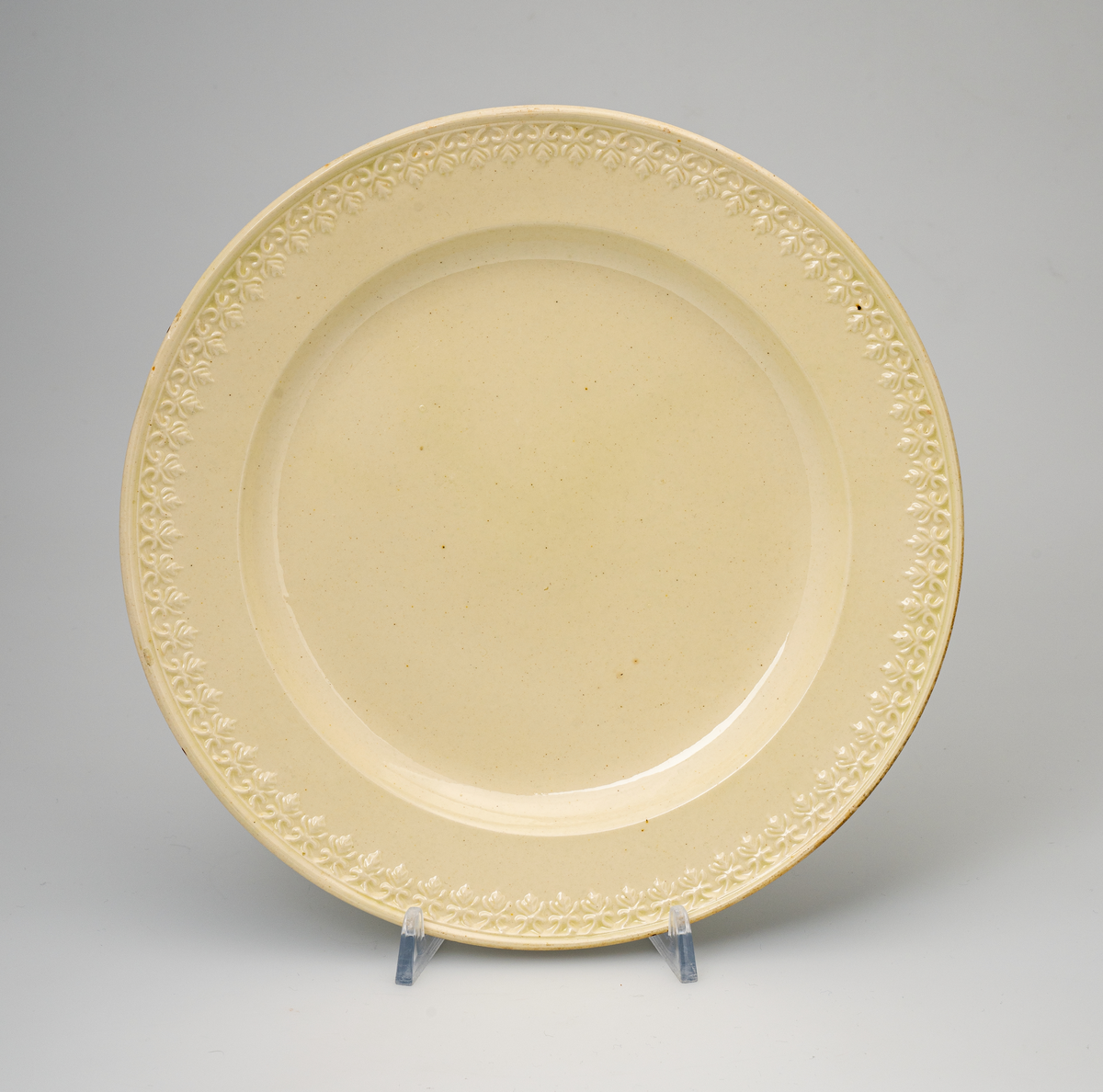 En flat tallerken av glasert steintøy (creamware). Den er hvitgul på farge (ensfarget). Flaten på midten er 16,5cm i diameter. Langs kanten er det tungekant. Innenfor tungekanten er det støpt dekor i form av en hjertebord med stiliserte blomster/bladverk. På undersiden er det dypstempel i form av fire små ruter/prikker i klase.
