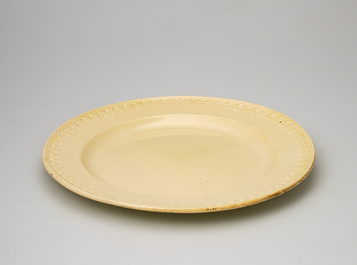 En flat tallerken av glasert steintøy (creamware). Den er hvitgul på farge (ensfarget). Flaten på midten er 16,5cm i diameter. Langs kanten er det tungekant. Innenfor tungekanten er det støpt dekor i form av en hjertebord med stiliserte blomster/bladverk. På undersiden er det dypstempel i form av fire små ruter/prikker i klase.
