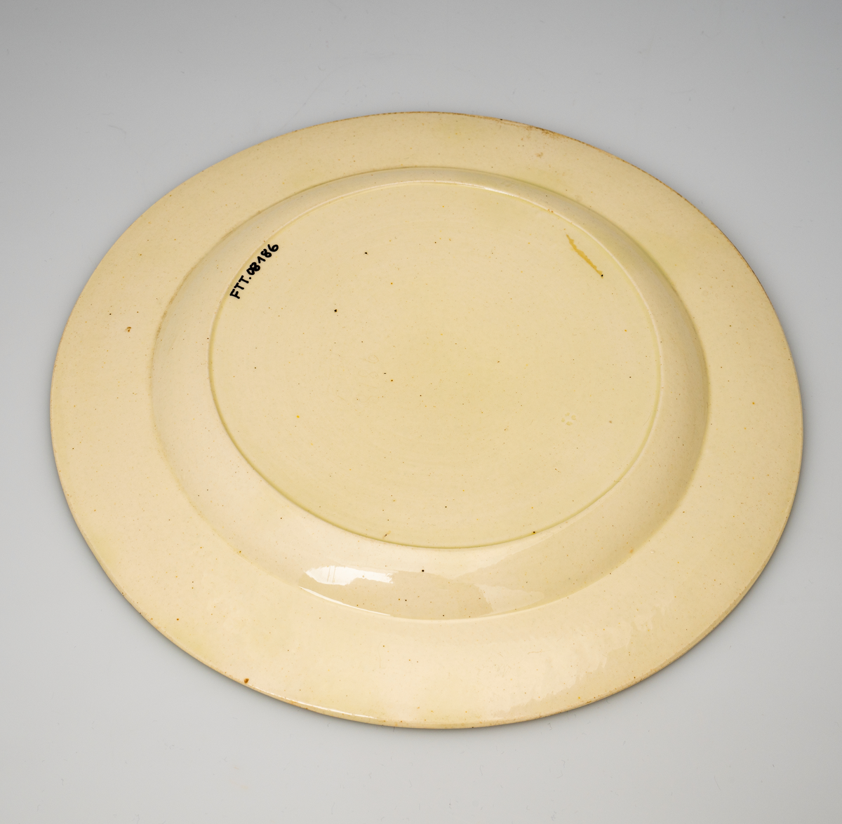 En flat tallerken av glasert steintøy (creamware). Den er hvitgul på farge (ensfarget). Flaten på midten er 16,5cm i diameter. Langs kanten er det tungekant. Innenfor tungekanten er det støpt dekor i form av en hjertebord med stiliserte blomster/bladverk. På undersiden er det dypstempel i form av fire små ruter/prikker i klase.
