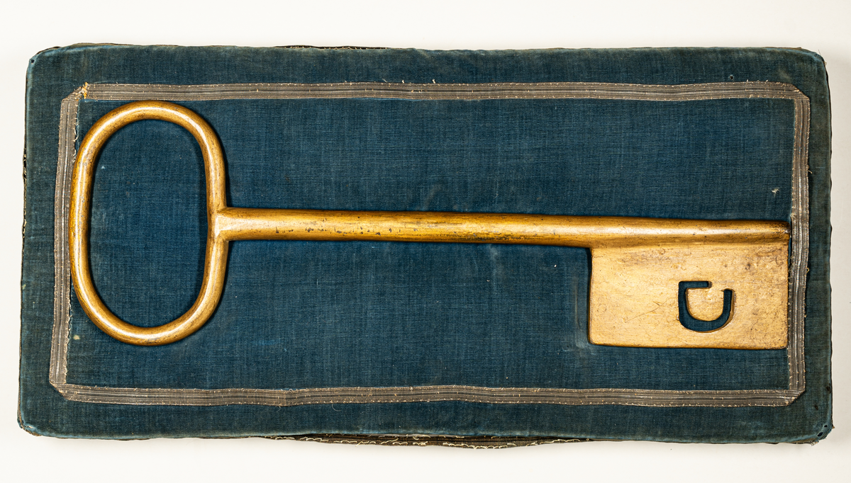 Stadens nyckel, förgyllt järn, genombrutet G i axet. Ligger på ett blått hyende (kudde, dyna) av sammet med silverbroderat band utmed sidorna.