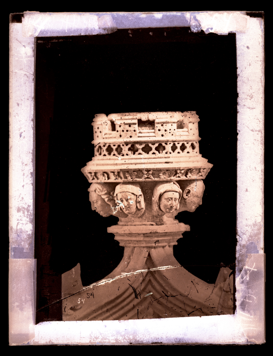 Kapitel på toppen av korbueveggen i Nidarosdomen, dekorert med åtte hoder. Konsoll for Kristusfigur. 
Bildet skal i følge Hovedkatalog for fotografier være tatt før restaurering. Korbueveggen ble restaurert i perioden 1871-1872.