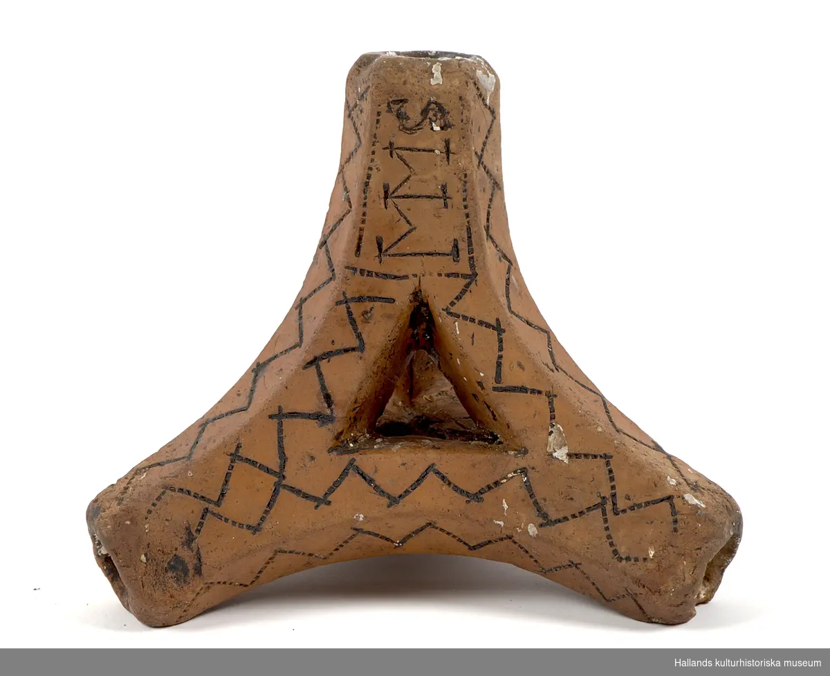 Ljusstake, en s.k, "Fotangelstake", av bränt lergods. Triangulär form, består av fyra pipor. Rikligt dekorerad med präglade sicksackmönster som förstärkts med sotning. I mönstret en signering: "MMS".